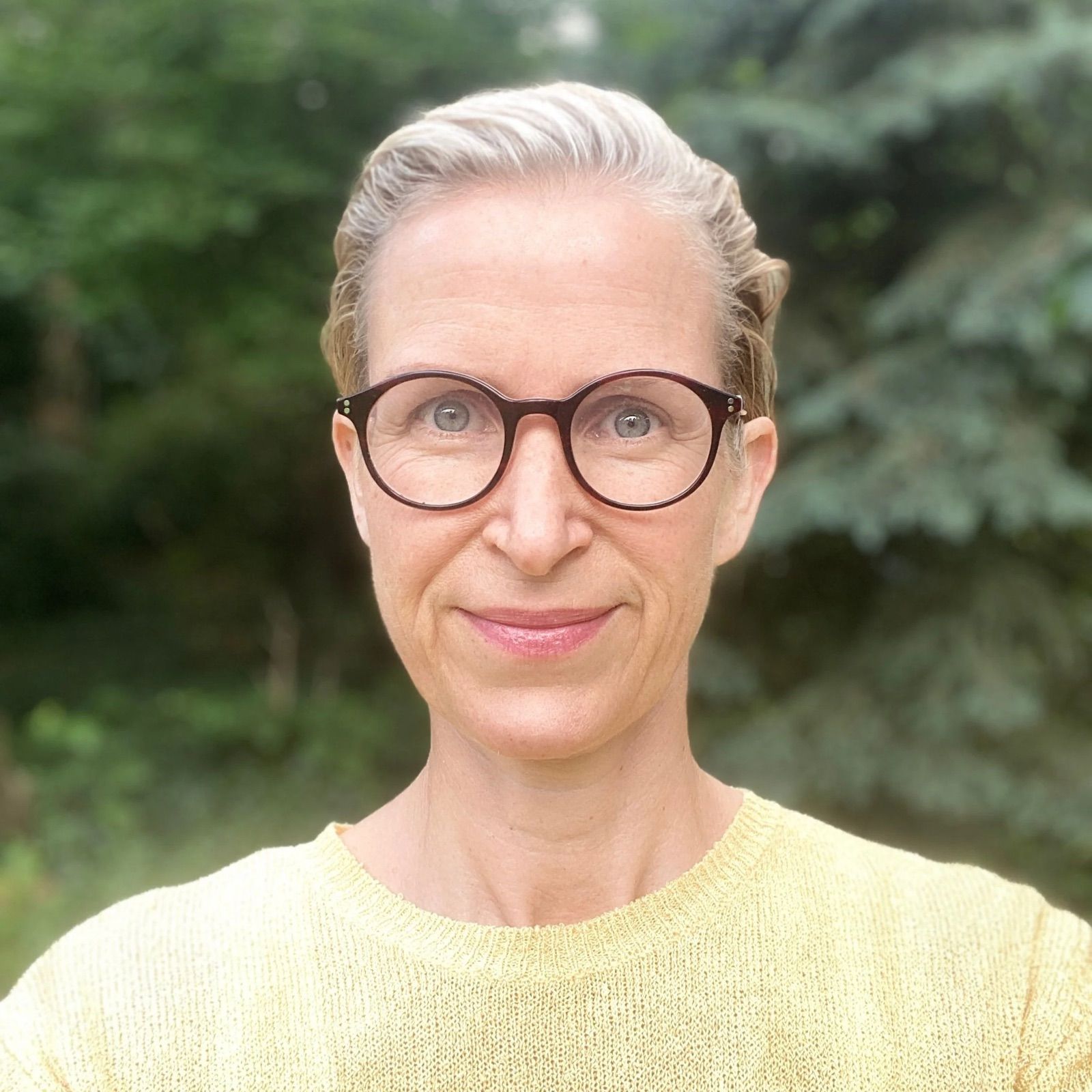 Portrait of a smiling older woman with glasses in front of a background of green trees.