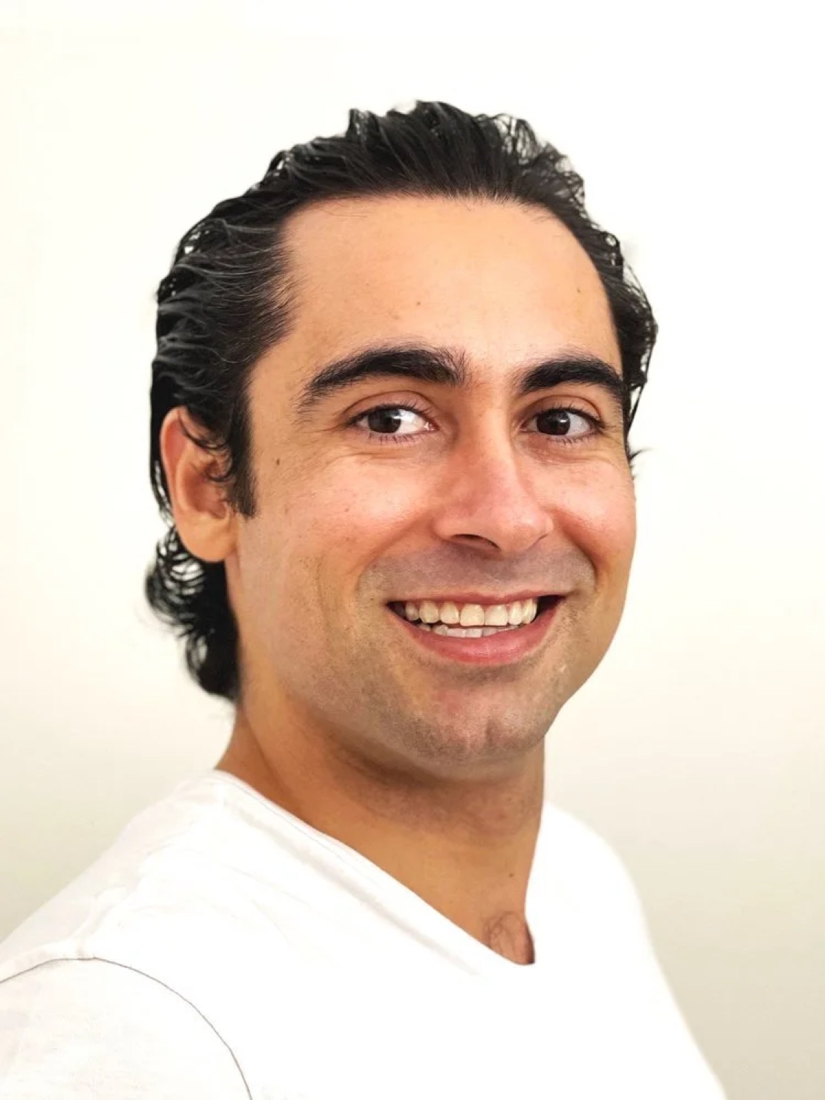 A man with dark, wavy hair, smiling, wearing a white T-shirt, in front of a light background.