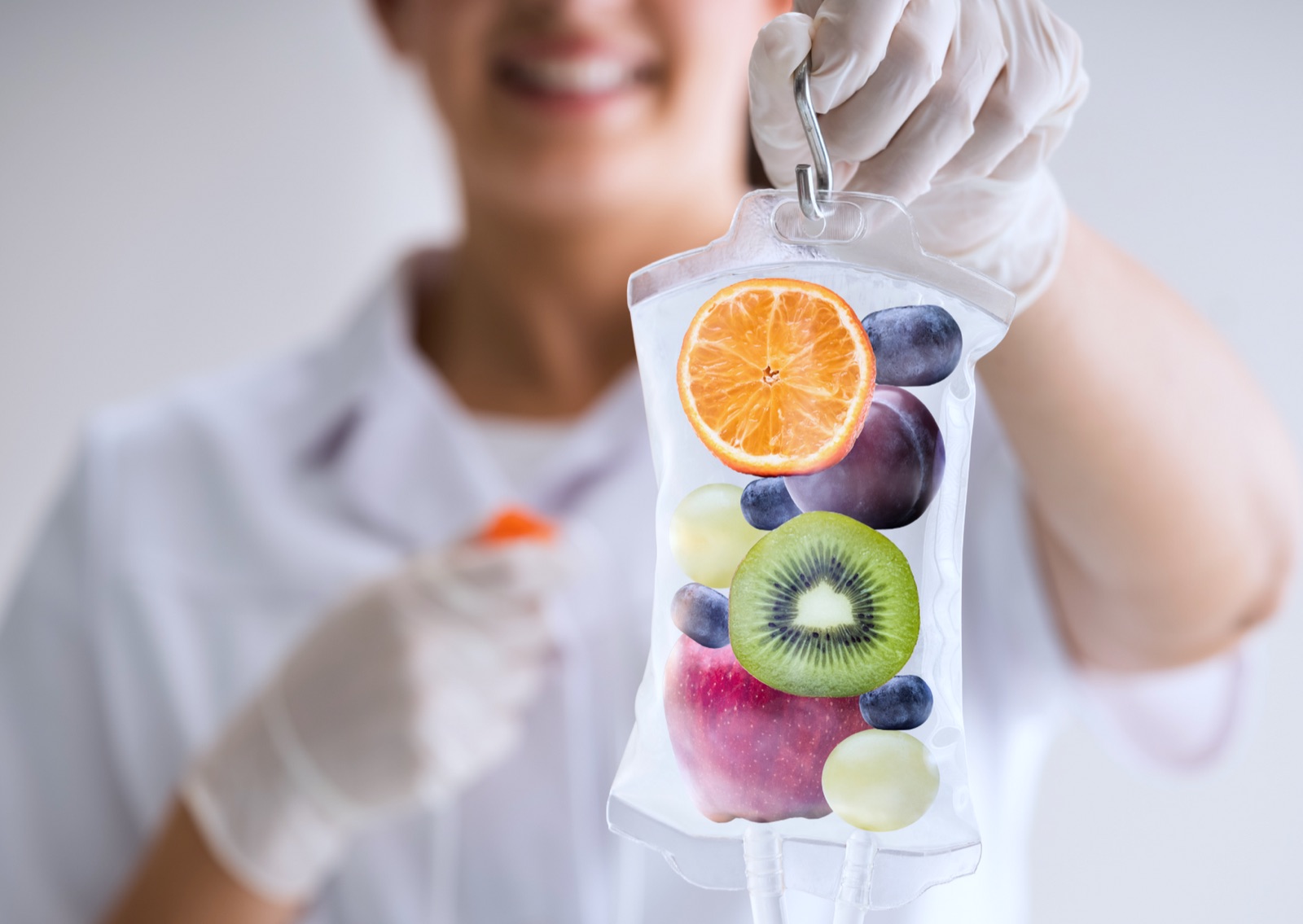 A person in white medical gloves holds a transparent medical fluid bag filled with fresh fruit such as orange, grapes, kiwi, apple and blueberries in front of the camera.