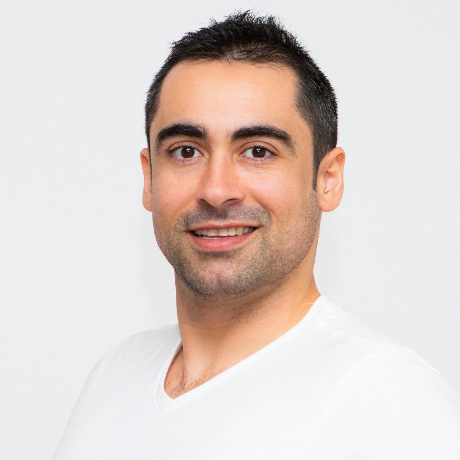 Portrait of a laughing man with dark hair, brown eyes and a light beard, wearing a white T-shirt, in front of a neutral background.