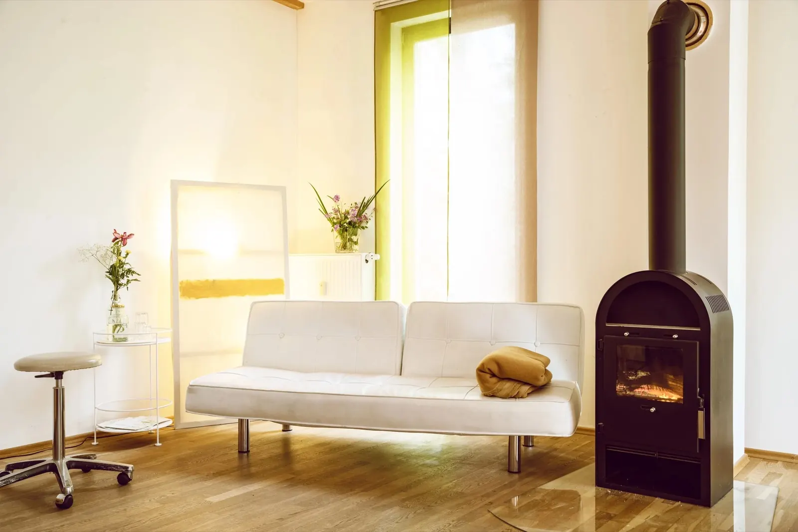 Gemütliches Wohnzimmer mit einem weißen Sofa, einem kleinen Beistelltisch mit Blumen, einem Heizkamin und einer großen Fensterfront mit grüner Vorhang.