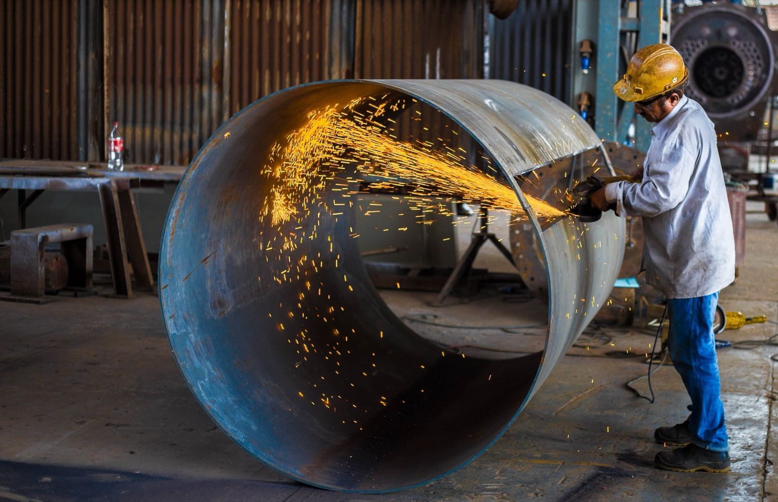 Worker welding a large metal pipe with flying sparks in a workshop.