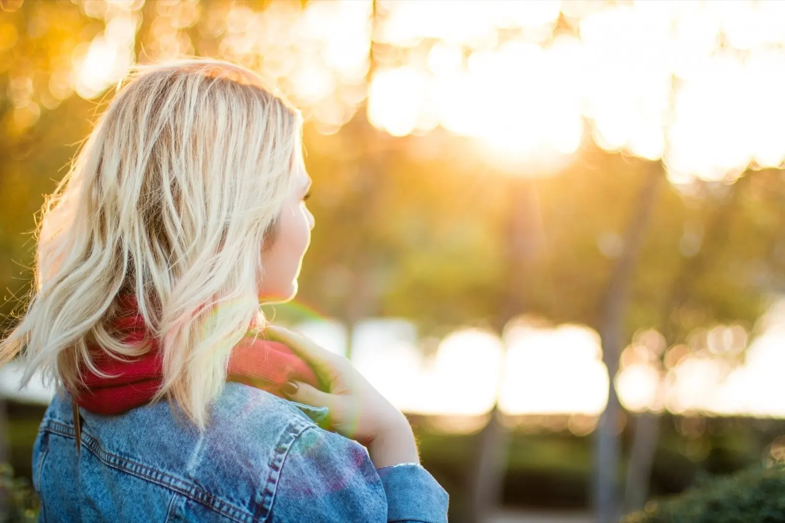 Frau mit blonden Haaren in einem grauen Mantel und roter Schals schaut in die Sonne in einem Park im Sonnenuntergang.