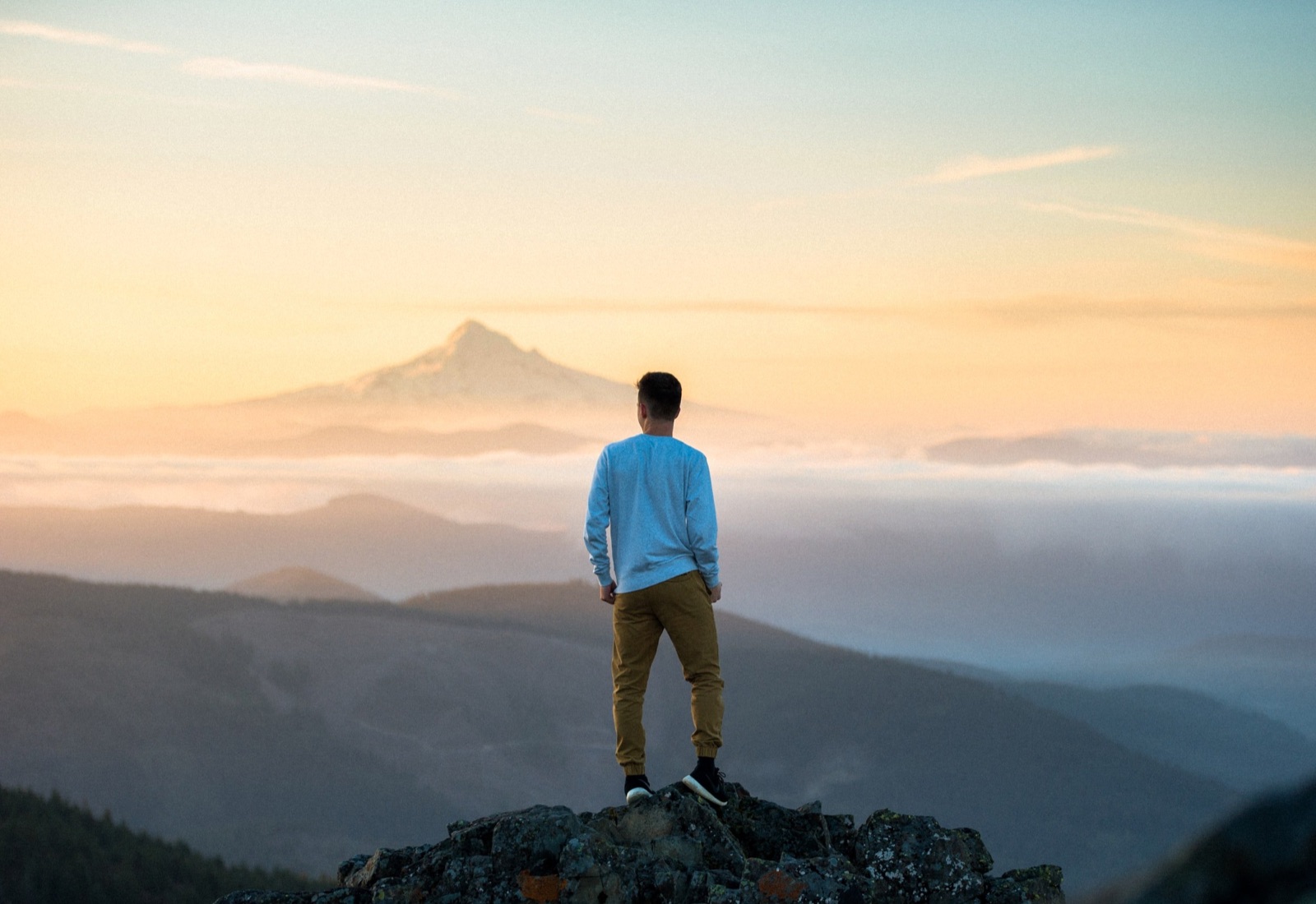 A young man stands on a rock looking at a mountain at sunrise or sunset, with clouds in the valley and a blue sky.