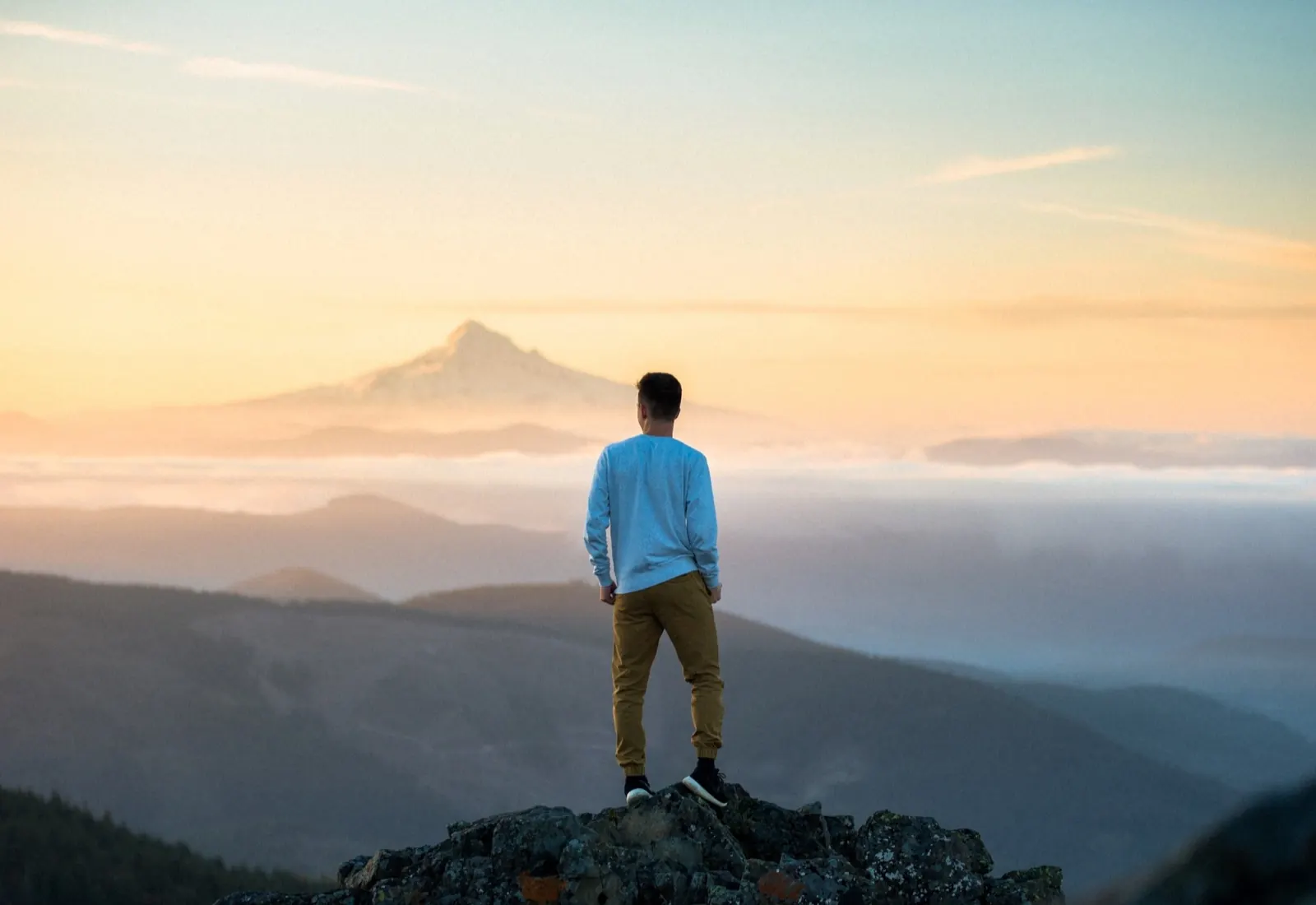 Ein junger Mann steht auf einem Felsen und blickt auf einen Berg im Sonnenaufgang oder Sonnenuntergang, mit Wolken im Tal und einem blauen Himmel.