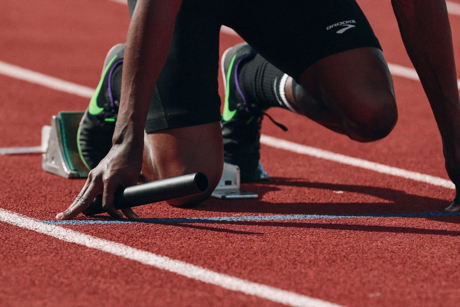 An athlete in starting position on a running track, ready to race, with starting blocks and a starting baton in hand.