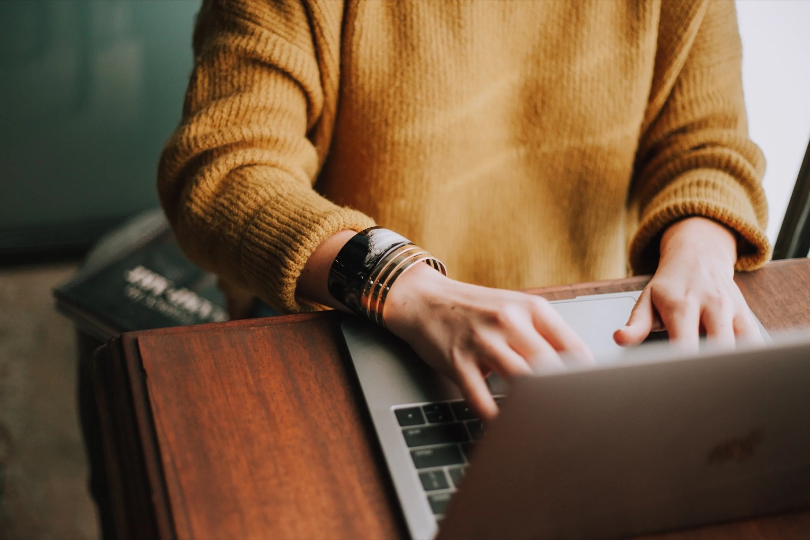 Close-up of a person typing on a laptop, wearing a large black and gold statement wristwatch and a brown sweater, sitting at a wooden table.
