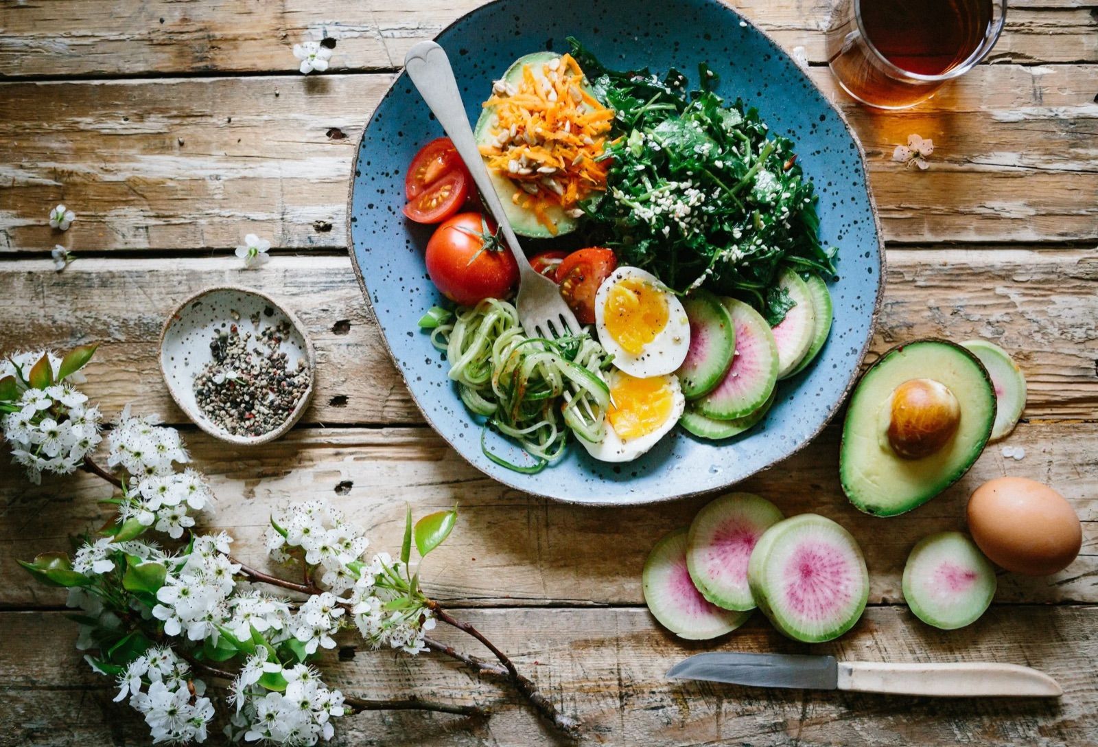 Vegetable salad with tomatoes, cucumbers, eggs, avocado, and red watercress, with a glass of tea on a wooden table, with pepper and salt, chives, a knife, flowers, and an egg