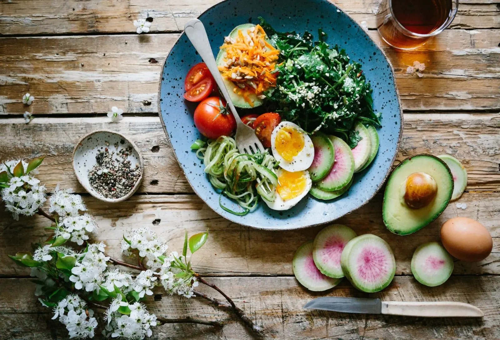 Bunte Salat mit Tomaten, Gurken, hartgekochten Eiern, Avocado und Karotten auf einem blauen Teller, daneben eine halbe Avocado, ein rohes Ei, Radieschen und ein Messer auf einem Holztisch. Es gibt auch ein Glas dunklen Getränks und eine kleine Schale mit Pfeffer. Blumen liegen auf dem Tisch.