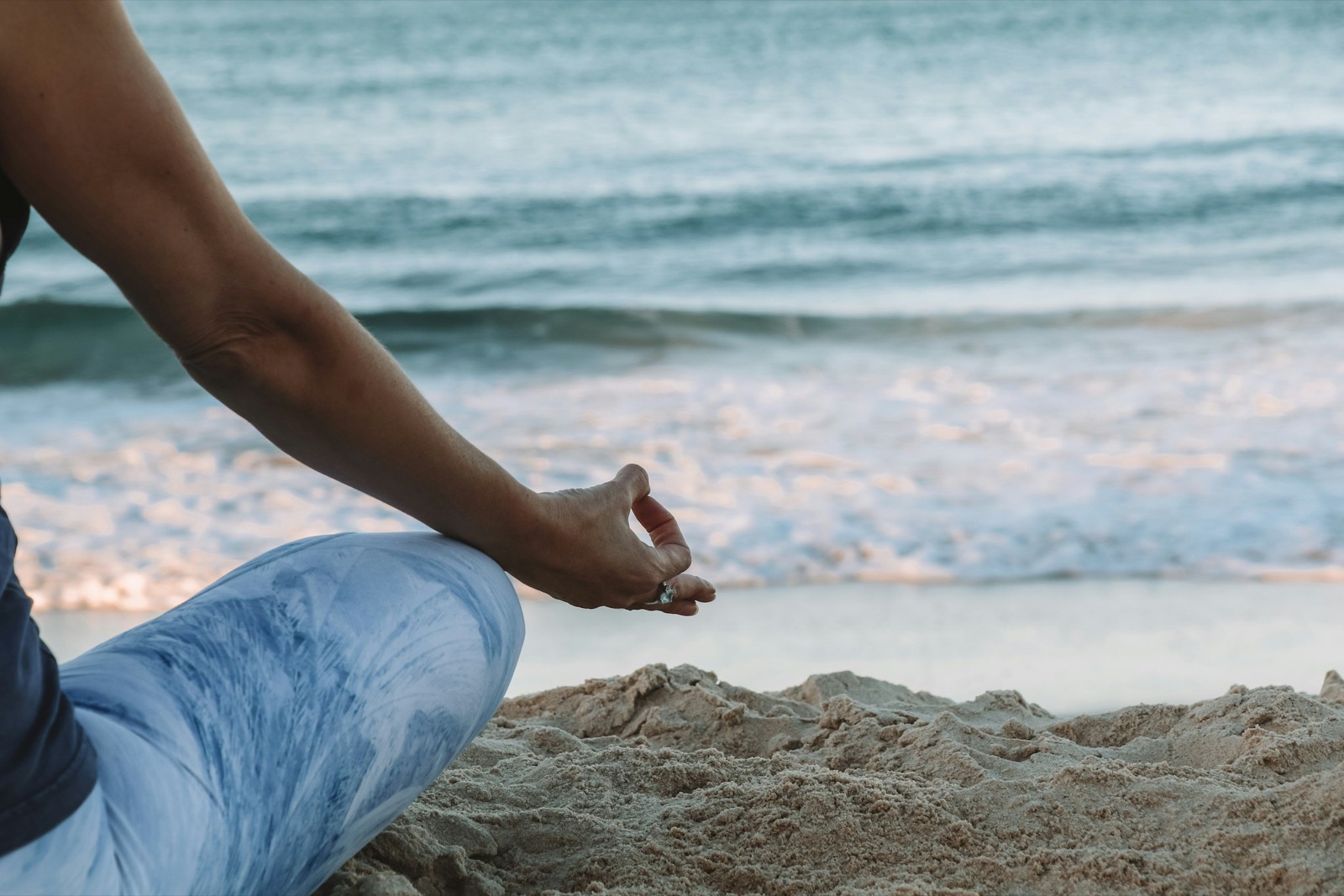 Person sitting cross-legged on the beach, holding one hand in Gyan Mudra, looking out at the sea.
