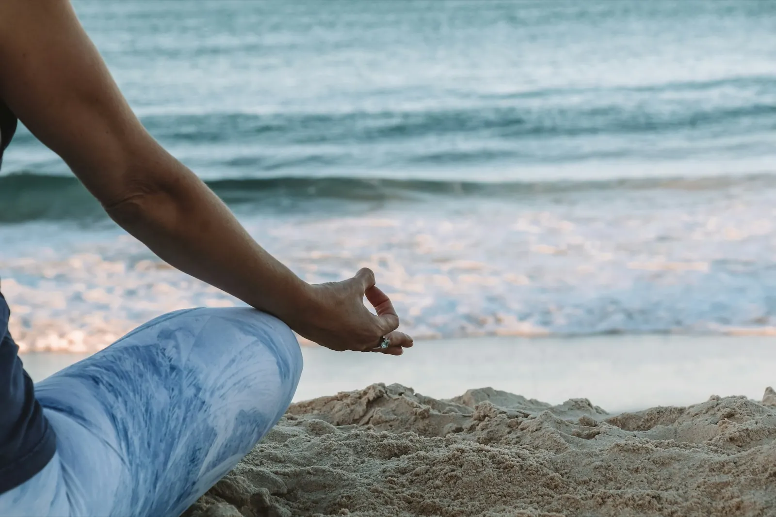 Person, die im Schneidersitz am Strand sitzt und eine Hand im Gyan Mudra hält, Blick auf das Meer.