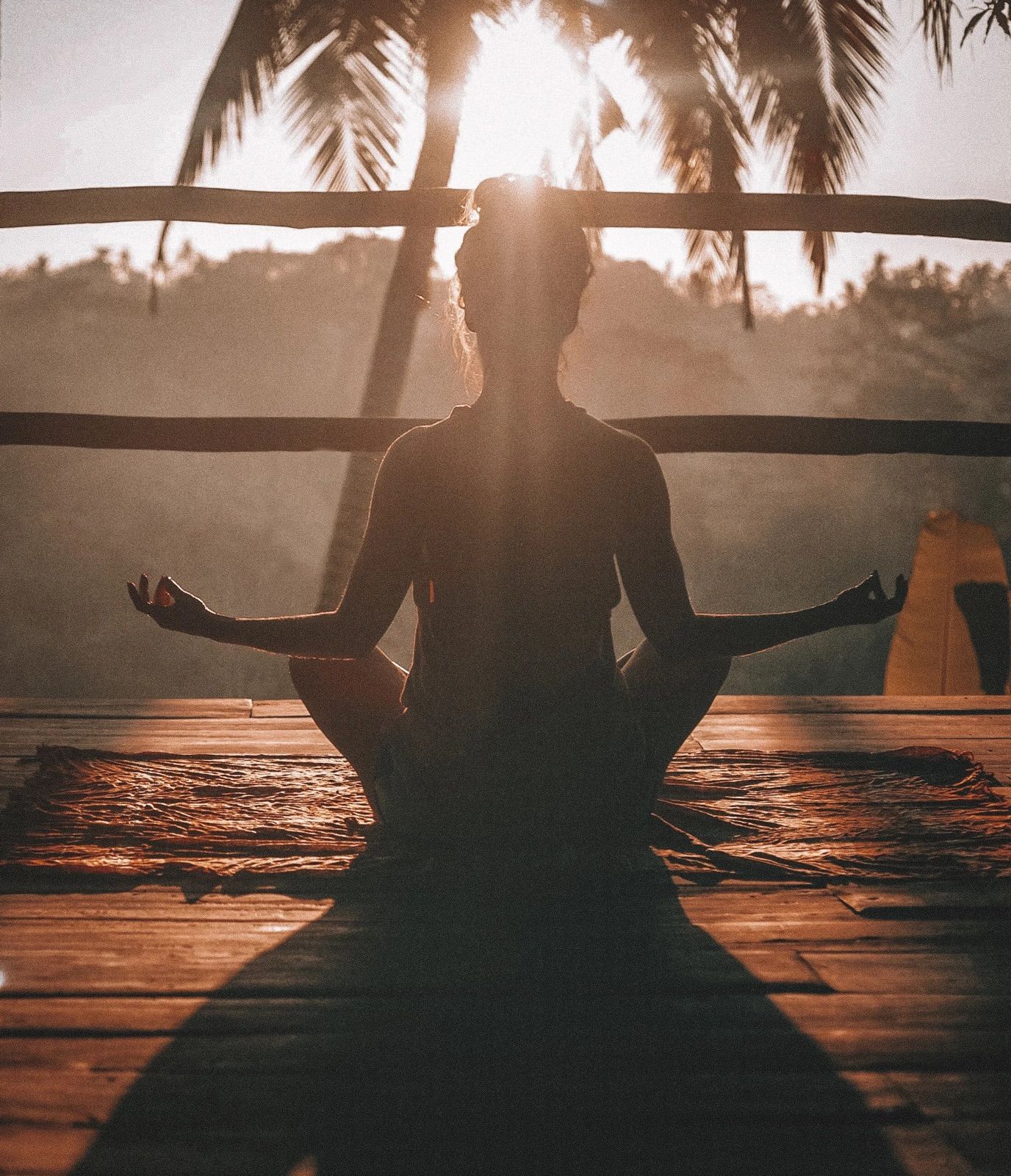 Person meditating in the lotus position on a wooden deck at sunset, surrounded by palm trees.