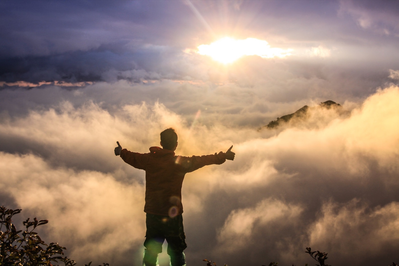 A person stands on a hill with outstretched arms in front of a sunrise or sunset, surrounded by clouds and mist over mountains.
