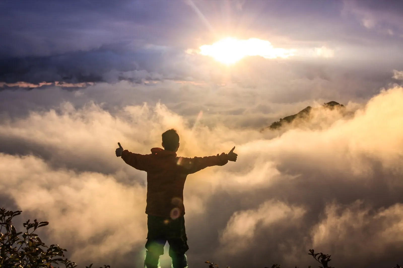 Ein Mensch steht auf einem Hügel, mit ausgestreckten Armen, vor einer Sonnenaufgangs- oder Sonnenuntergangsszene, umgeben von Wolken und Nebel über Bergen.